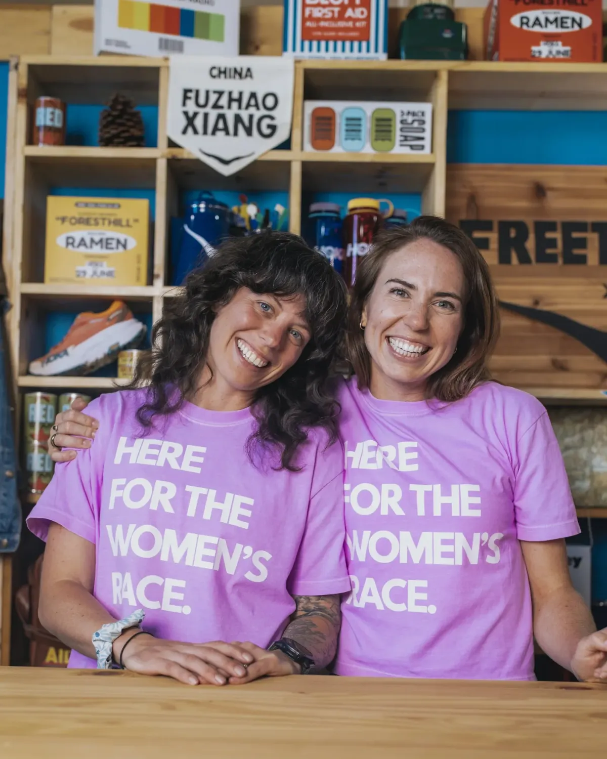 Two women in pink Here for the Women's Race tees, grinning, at a trail race expo.