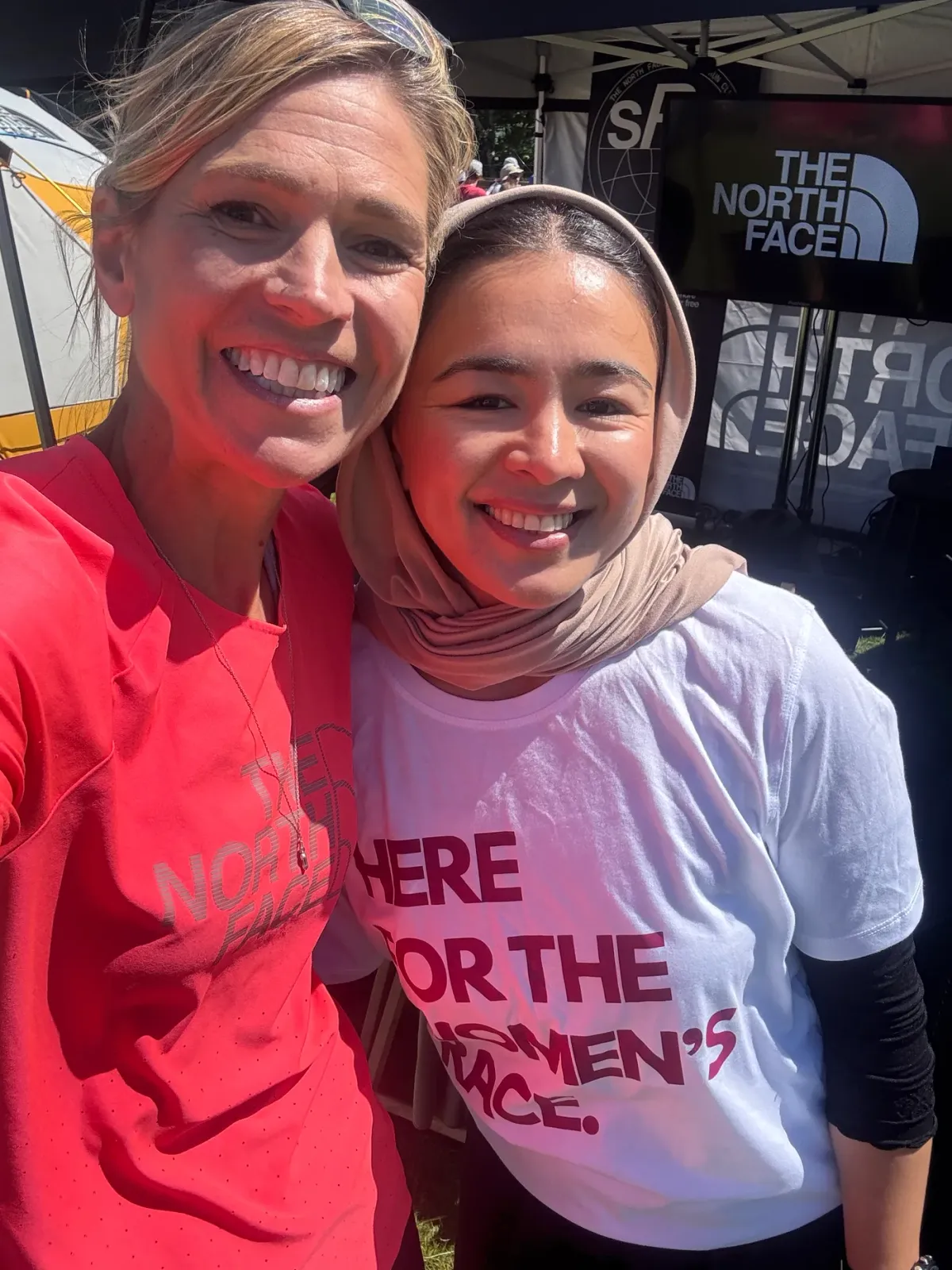 Two runners, one in a headscarf and the Here for the Women's Race tee, smiling at a race village.