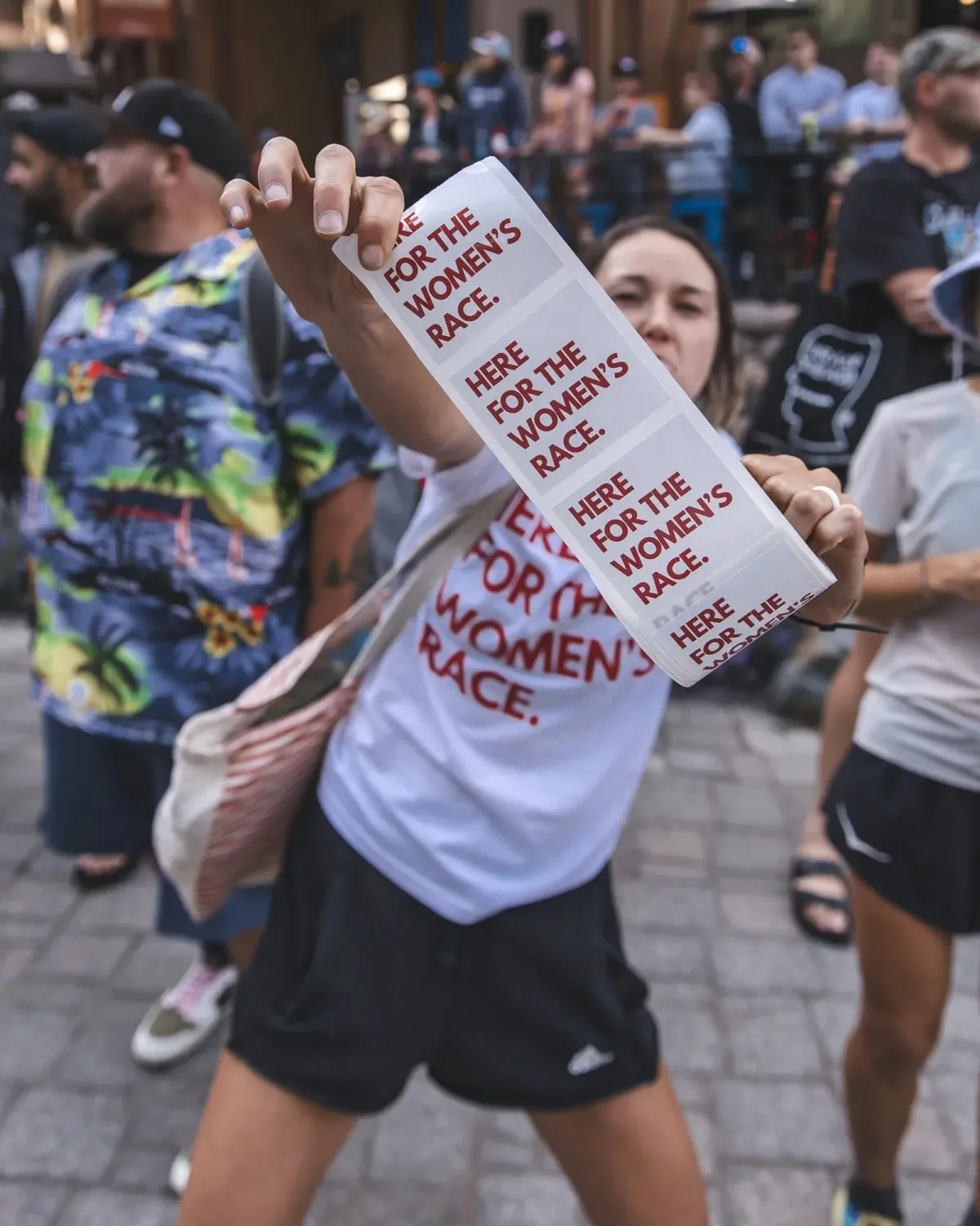 A woman at a race expo holds up a roll of Here for the Women's Race stickers, grinning, wearing the tee.